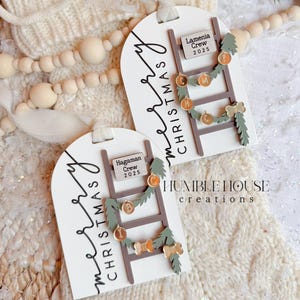 May include: Two white Christmas gift tags with a ladder design, garland, and the words "Merry Christmas." Each tag has a small sign with a name and the year 2025. The tags are decorated with gold and green accents.