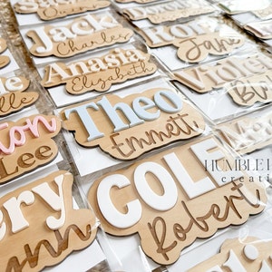 May include: A collection of personalized wooden name signs with various names, including Theo, Emmett, Cole, and Robert. The names are painted in white, pink, and blue on a natural wood background. The signs are packaged in clear plastic bags. The image also includes the text "HUMBLE HOUSE creations".