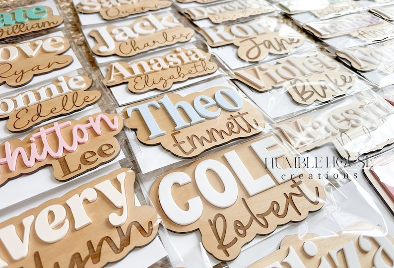 May include: A collection of personalized wooden name signs with various names, including Theo, Emmett, Cole, and Robert. The names are painted in white, pink, and blue on a natural wood background. The signs are packaged in clear plastic bags. The image also includes the text "HUMBLE HOUSE creations".