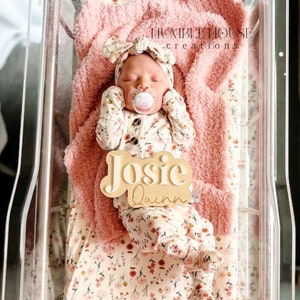 May include: A newborn baby girl wearing a floral print onesie and a pink bow headband is lying in a clear plastic bassinet. A wooden name sign with the names "Josie Quinn" is placed next to the baby.