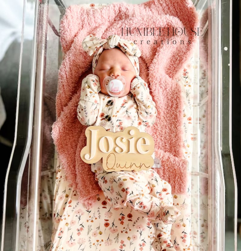 May include: A newborn baby girl wearing a floral print onesie and a pink bow headband is lying in a clear plastic bassinet. A wooden name sign with the names "Josie Quinn" is placed next to the baby.