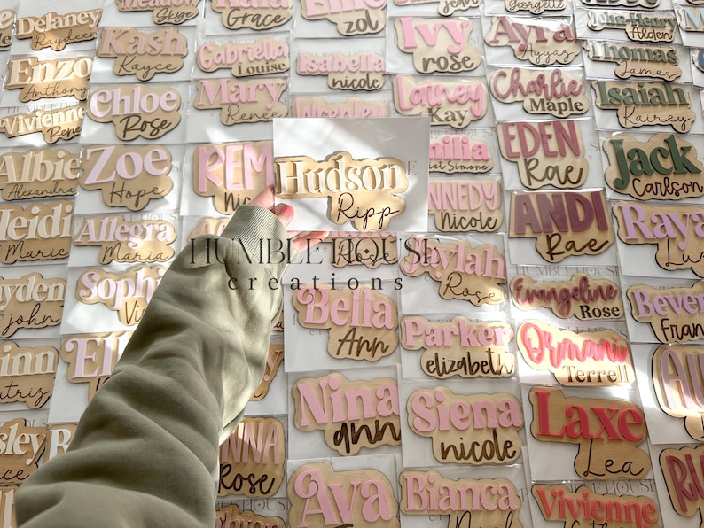 May include: A collection of personalized wooden name tags with various names written in pink and white lettering. The names are displayed on a white background with a light brown border.