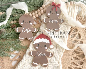 May include: Three gingerbread person ornaments with personalized names. The ornaments are brown with white icing and are decorated with red and white accents. The names on the ornaments are Cole, Hazel, and Cohen.