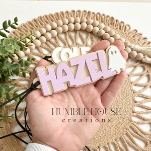May include: A purple wooden name tag with the name "HAZEL" and a white ghost cutout. The name tag is attached to a black cord.