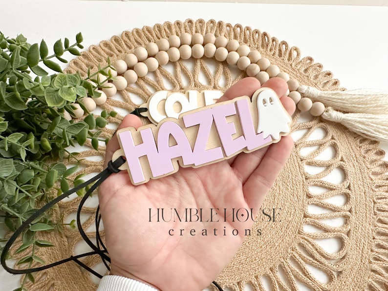 May include: A purple wooden name tag with the name "HAZEL" and a white ghost cutout. The name tag is attached to a black cord.