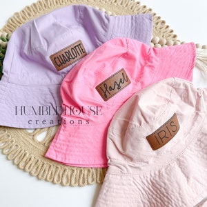 May include: Three sun hats in pink, light pink, and purple with brown leather patches that say "Charlotte", "Hazel", and "Iris".