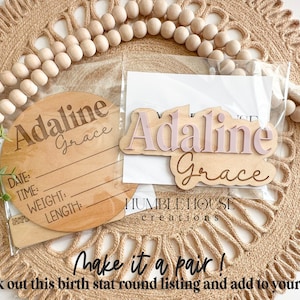 May include: A set of two wooden name plaques with the name "Adaline Grace" in pink and brown lettering. The larger plaque is a round birth stat plaque with lines for date, time, weight, and length. The smaller plaque is a rectangular name plaque. The plaques are packaged together in a clear plastic bag.