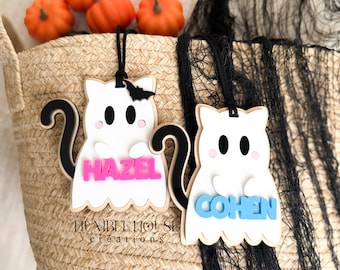 May include: Two white ghost-shaped wooden ornaments with black tails and hanging cords. One ghost features the pink name HAZEL, and the other features the blue name COHEN. Both ghosts have black eyes, pink cheeks, and small white arms. A small black bat is attached to the top of the first ghost.