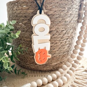 May include: A wooden Halloween door hanger with the word "Cole" in white letters and a carved orange pumpkin with a jack-o-lantern face. The hanger is attached to a black string and is hanging from a wicker basket.
