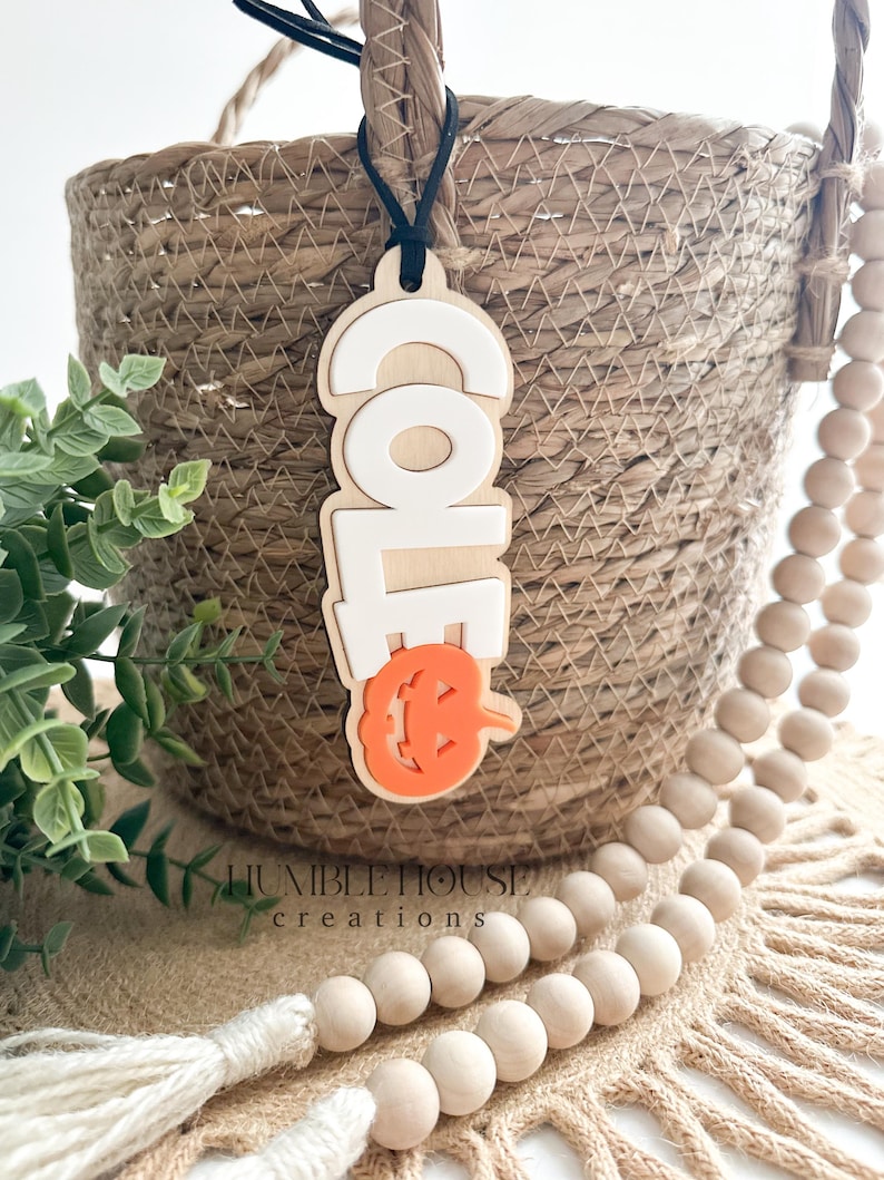 May include: A wooden Halloween door hanger with the word "Cole" in white letters and a carved orange pumpkin with a jack-o-lantern face. The hanger is attached to a black string and is hanging from a wicker basket.