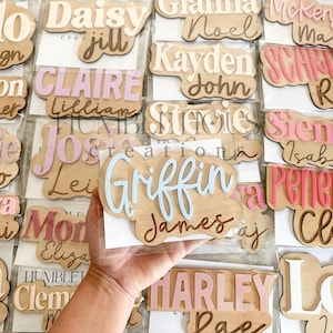 May include: A collection of wooden name plaques with various names written in different colors and fonts. The plaques are arranged in rows and are displayed on a white background.