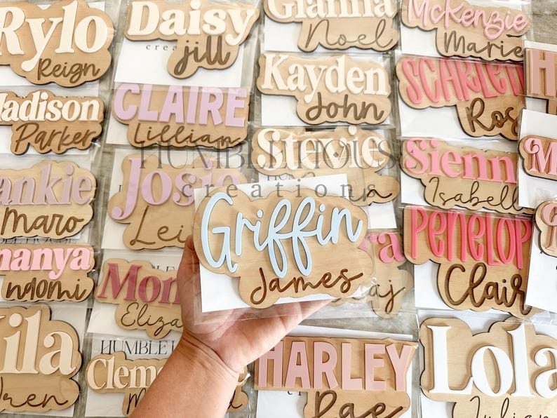May include: A collection of wooden name plaques with various names written in different colors and fonts. The plaques are arranged in rows and are displayed on a white background.
