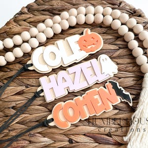 May include: Three wooden Halloween name tags with black string. The names are Cole, Hazel, and Cohen. Each tag has a Halloween-themed cutout: a pumpkin, a ghost, and a bat.