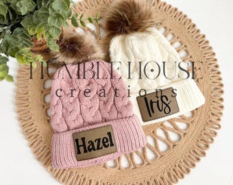 Peut inclure: Deux bonnets d'hiver en tricot avec des pompons en fausse fourrure. Le bonnet rose a un patch en cuir marron avec le nom "Hazel" dessus. Le bonnet blanc a un patch en cuir marron avec le nom "Iris" dessus.