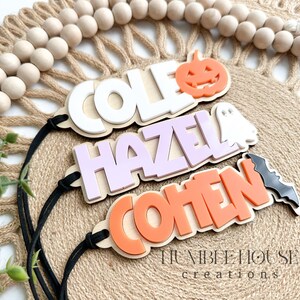 May include: Three Halloween-themed wooden name tags with black string. The top tag is white with the name "COLE" and a pumpkin cutout. The middle tag is purple with the name "HAZEL" and a ghost cutout. The bottom tag is orange with the name "COHEN" and a bat cutout.