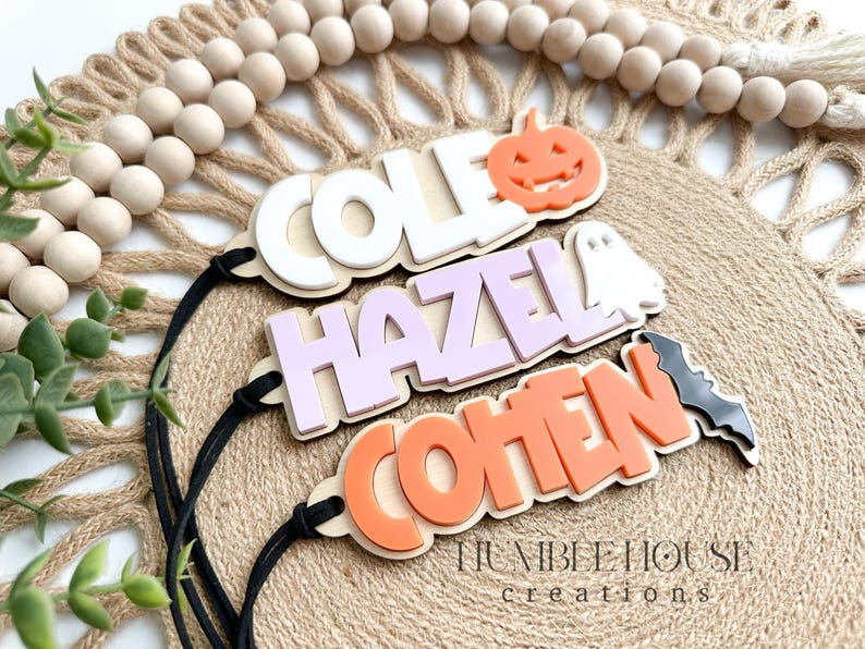 May include: Three Halloween-themed wooden name tags with black string. The top tag is white with the name "COLE" and a pumpkin cutout. The middle tag is purple with the name "HAZEL" and a ghost cutout. The bottom tag is orange with the name "COHEN" and a bat cutout.