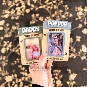 May include: Two wooden photo frames, one labeled "DADDY" and the other "POPPOP", each with a photo of a child. The frames have a light brown background with green and blue accents and the text "YOU'RE THE BEST!" and "HAPPY FATHERS DAY 2026".