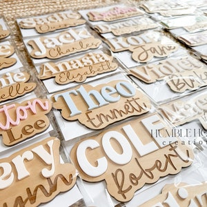 May include: A collection of personalized wooden name plaques with various names written in different colors and fonts. The plaques are displayed on a brown surface and are packaged in clear plastic bags. The text "HUMBLE HOUSE creations" is visible in the bottom right corner of the image.