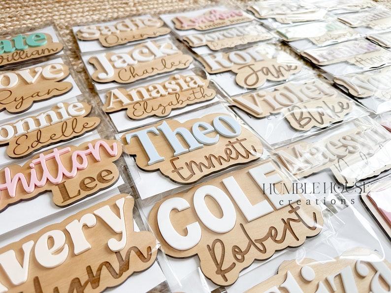 May include: A collection of personalized wooden name plaques with various names written in different colors and fonts. The plaques are displayed on a brown surface and are packaged in clear plastic bags. The text "HUMBLE HOUSE creations" is visible in the bottom right corner of the image.