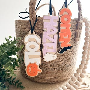 May include: Three Halloween-themed wooden name tags with the names "Cole", "Hazel", and "Cohen" in different colors. The tags are hanging from black cords and are in a wicker basket with a wooden bead garland.