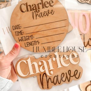 May include: A set of wooden baby name plaques with various names and fonts. The plaques are in different shapes and sizes, and some have a space for birth information. The plaques are in a variety of colors, including pink, blue, and white.