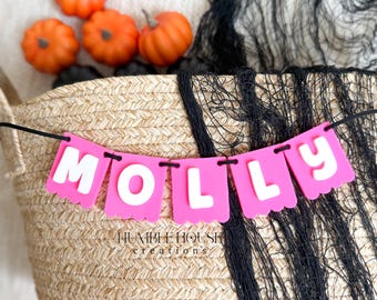May include: A pink banner with white letters spelling MOLLY strung on a black cord, displayed in front of a woven basket containing orange pumpkins and black decorative netting. The text HUMBLE HOUSE creations is visible on the basket.