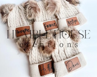 Peut inclure: Cinq bonnets en tricot crème avec des pompons en fausse fourrure. Chaque bonnet a un patch en cuir marron avec un nom écrit en noir : Amarah Rae, Emmie, Westlynn, Hattie et Demi.