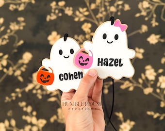 May include: Two white ghost-shaped ornaments with black outlines and black hanging cords. One ghost features the name Cohen in black text with an orange pumpkin bucket, while the other features the name Hazel with a pink bow and pink pumpkin bucket.