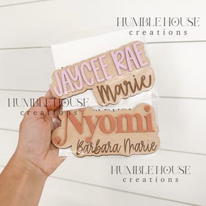 May include: Three wooden name plaques with pink and orange lettering. The top plaque reads "Jaycee Rae Marie", the middle plaque reads "Nyomi", and the bottom plaque reads "Barbara Marie".