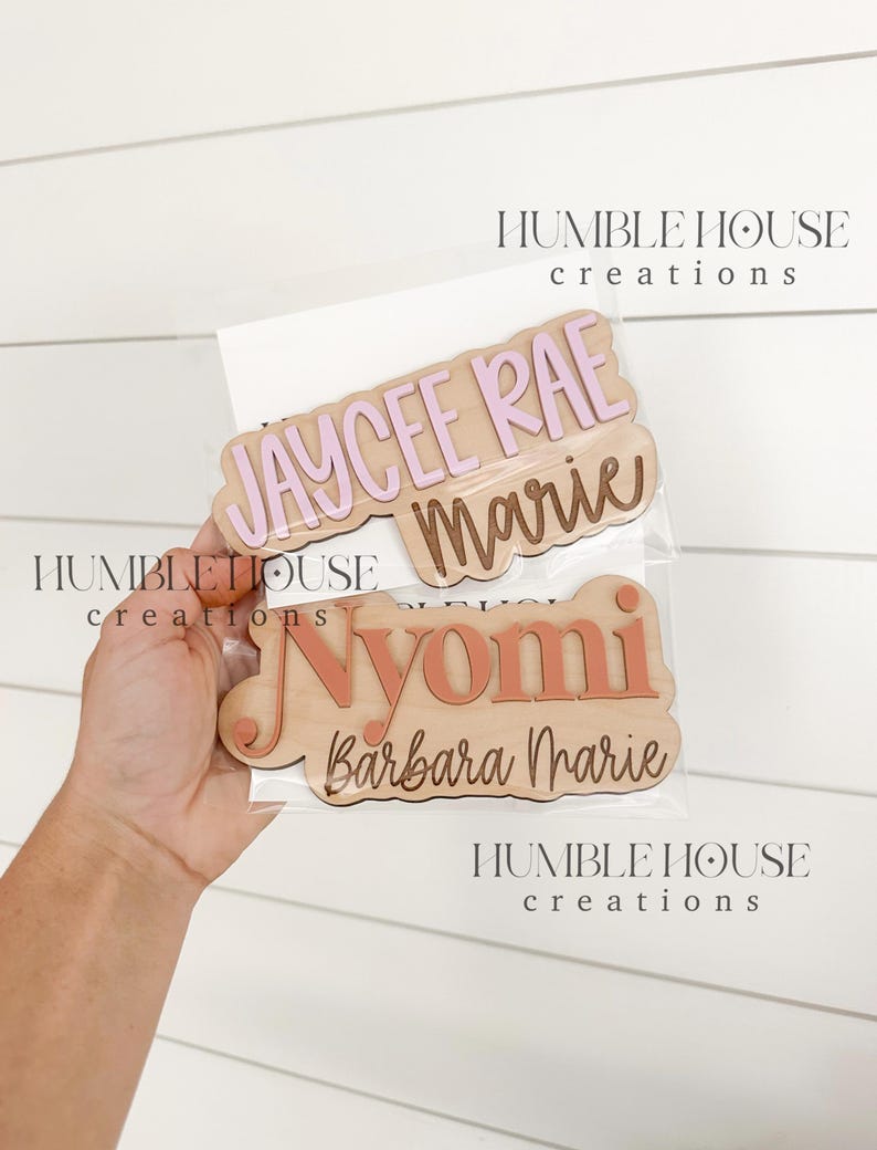 May include: Three wooden name plaques with pink and orange lettering. The top plaque reads "Jaycee Rae Marie", the middle plaque reads "Nyomi", and the bottom plaque reads "Barbara Marie".