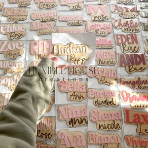 May include: A collection of personalized wooden name tags with various names written in pink and white lettering. The names are displayed on a white background with a light brown border.