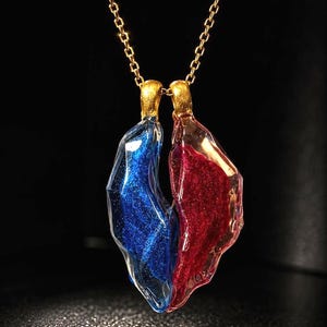 Puede incluir: Un collar de cadena dorada con dos colgantes. Un colgante es azul con purpurina, el otro es rojo con purpurina. Los colgantes están unidos en la parte superior con metal dorado.