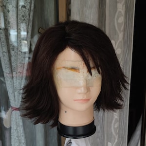 Puede incluir: Una cabeza de maniquí con una peluca corta de color marrón oscuro. La peluca tiene un estilo en capas. La cabeza del maniquí está sobre un soporte negro, con un paño blanco colgando debajo. El fondo incluye una ventana y cortinas.