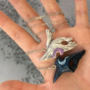 May include: Two silver chain necklaces with decorative pendants. One pendant is a white and purple mask with a detailed design. The other pendant is a black and blue mask with a detailed design.