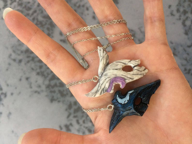 May include: Two silver chain necklaces with decorative pendants. One pendant is a white and purple mask with a detailed design. The other pendant is a black and blue mask with a detailed design.