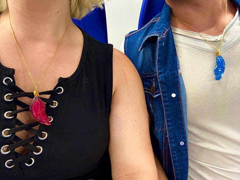 May include: Two gold chain necklaces with pendant charms. One pendant is a red, leaf-shaped charm, and the other is a blue, teardrop-shaped charm. The necklaces are worn over black and white tops.