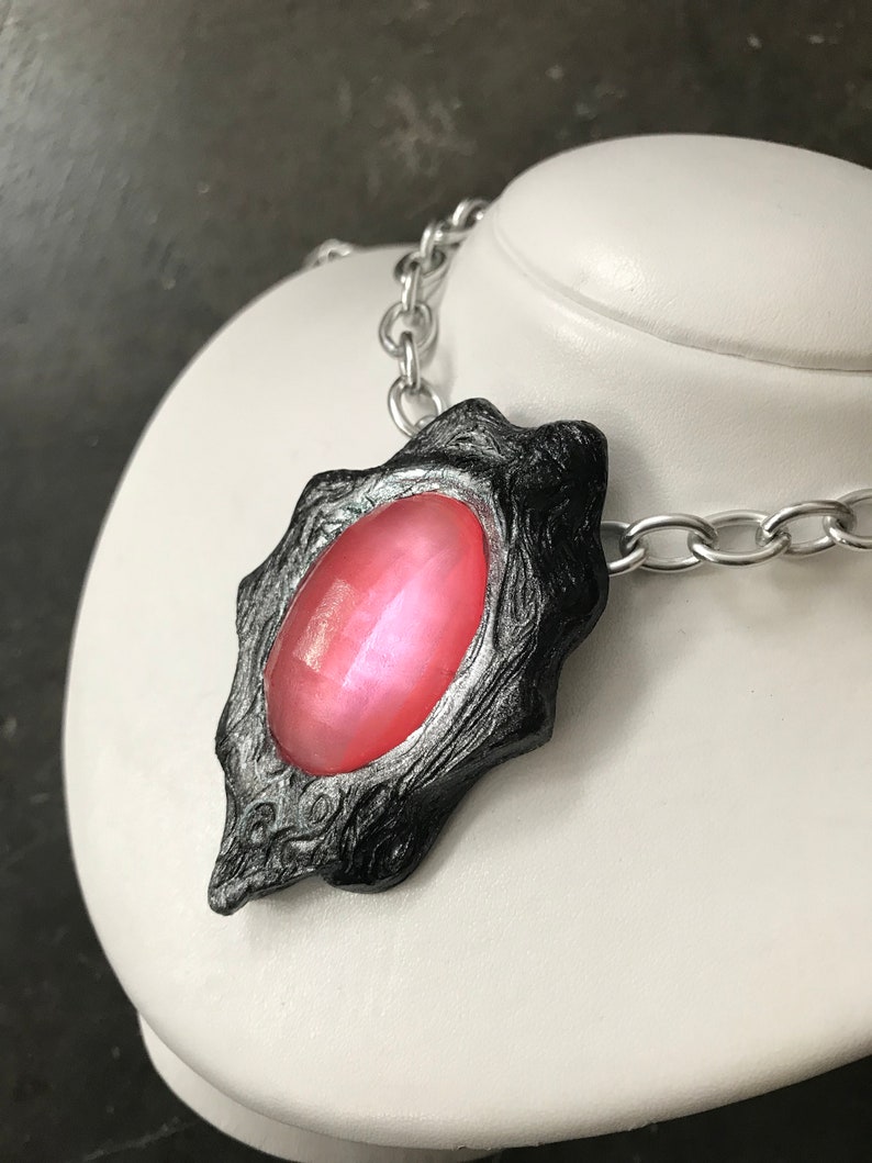 Pode incluir: Um colar de corrente prateada com um pingente preto e prateado com uma gema rosa. O pingente tem um design detalhado e texturizado.