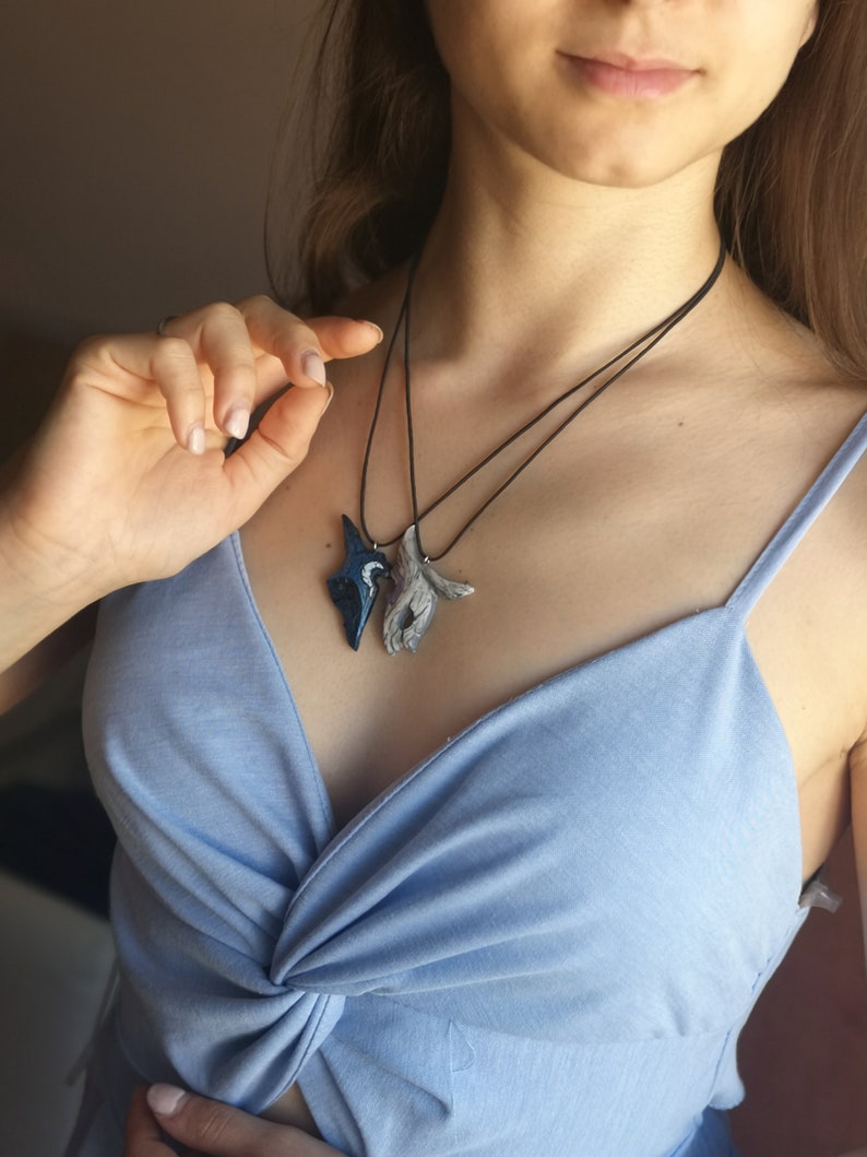 May include: A woman wearing a light blue top with a knotted front, is wearing two necklaces. One necklace features a blue and white bird pendant, and the other necklace features a pair of hands pendant.