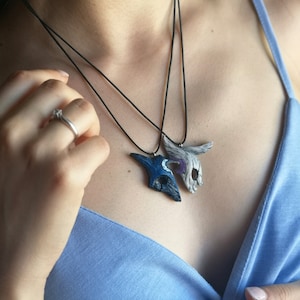 May include: Two necklaces with pendants shaped like masks. The masks are made of white and blue clay and have intricate details. The necklaces are black and are worn by a person.