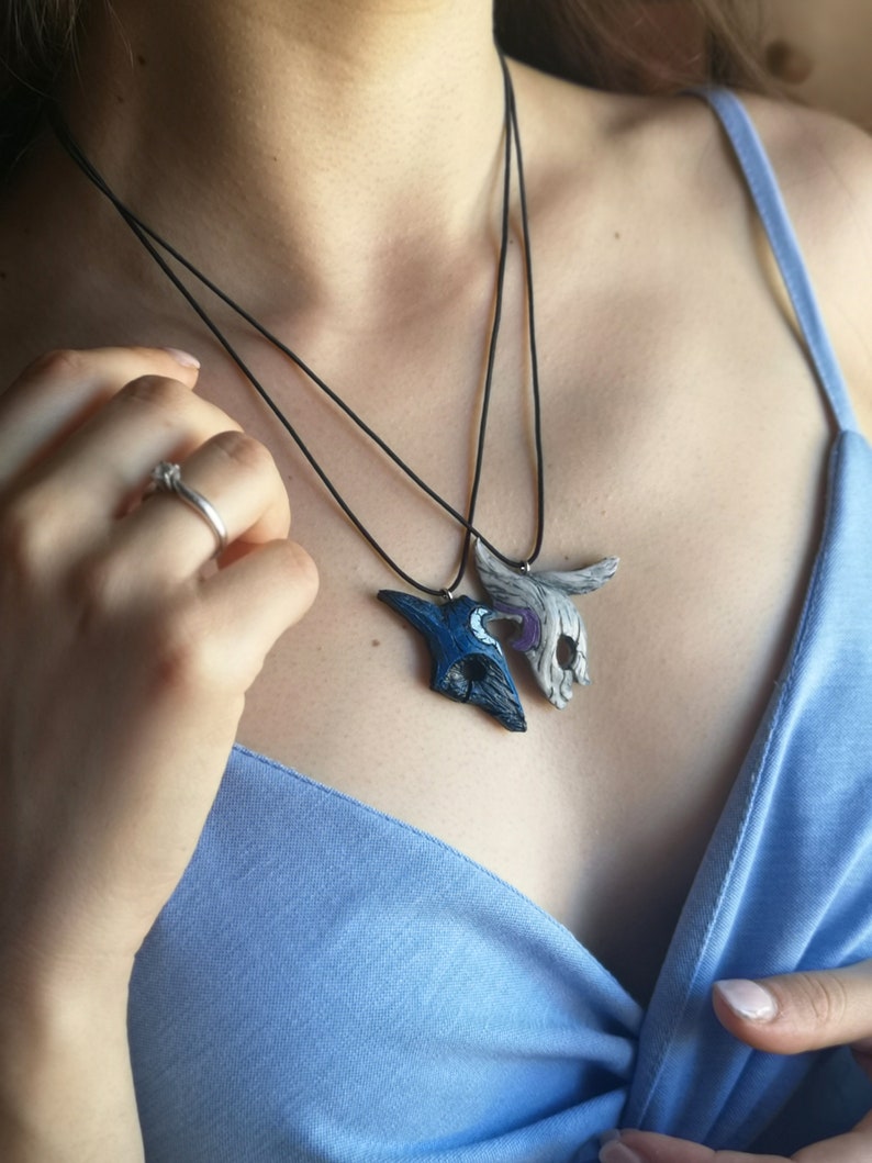 May include: Two necklaces with pendants shaped like masks. The masks are made of white and blue clay and have intricate details. The necklaces are black and are worn by a person.