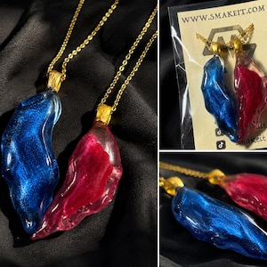 May include: Two resin pendants, one blue and one red, hang from gold chains. The pendants have an organic shape and a shimmering, glittery effect. The pendants are displayed on a black background. The brand name is visible on the packaging.