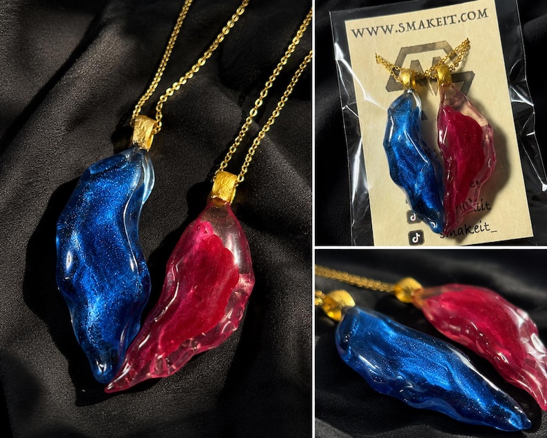 May include: Two resin pendants, one blue and one red, hang from gold chains. The pendants have an organic shape and a shimmering, glittery effect. The pendants are displayed on a black background. The brand name is visible on the packaging.