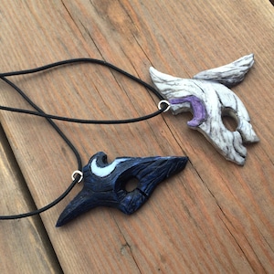 May include: Two black cord necklaces with pendants shaped like masks. One mask is white with purple accents and the other is black with white accents.