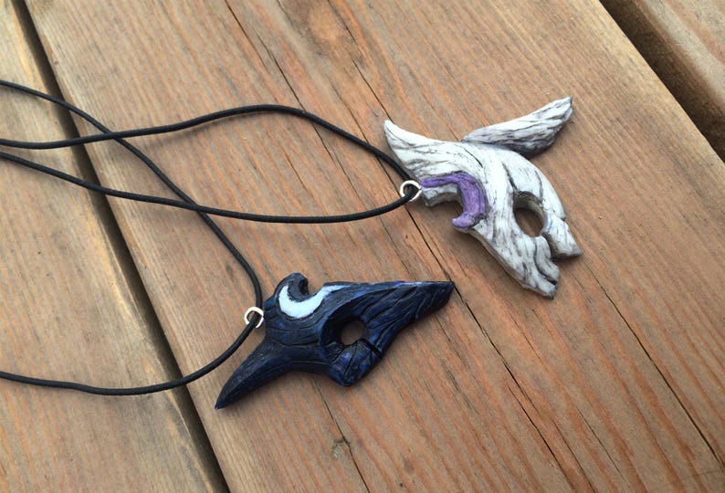 May include: Two black cord necklaces with pendants shaped like masks. One mask is white with purple accents and the other is black with white accents.
