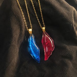 May include: Two necklaces with gold chains. Each necklace has a pendant in the shape of a jagged crystal. One pendant is blue, the other is red. The pendants have a glittery, shimmering effect.