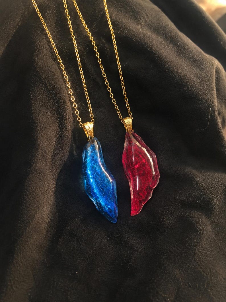 May include: Two necklaces with gold chains. Each necklace has a pendant in the shape of a jagged crystal. One pendant is blue, the other is red. The pendants have a glittery, shimmering effect.
