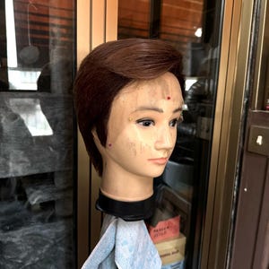 Może przedstawiać: Ruda peruka na głowie manekina z niebieskim ręcznikiem przewieszonym przez stojak. Peruka jest ułożona w krótkim, warstwowym cięciu.