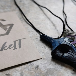 May include: A blue and white wooden pendant necklace with a black cord. The pendant is shaped like a bird's head with a crescent moon on the side. The necklace is hanging on a white surface with a brown paper tag that says "Make It" in black cursive.