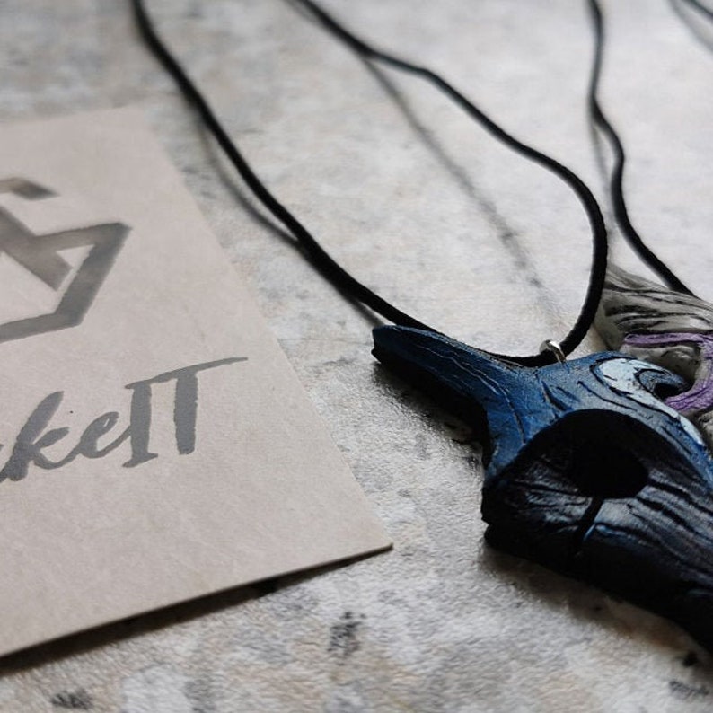 May include: A blue and white wooden pendant necklace with a black cord. The pendant is shaped like a bird's head with a crescent moon on the side. The necklace is hanging on a white surface with a brown paper tag that says "Make It" in black cursive.