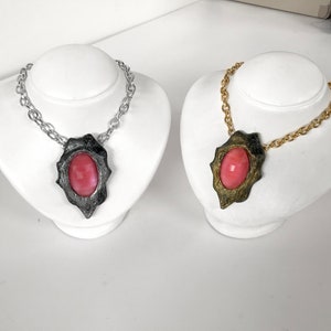 Pode incluir: Dois colares com pingentes de pedra rosa em expositores brancos. Um tem uma corrente prateada e o outro uma corrente dourada.